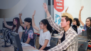 Sekelompok mahasiswa di kelas komputer sedang mengangkat tangan untuk menjawab pertanyaan, menunjukkan antusiasme dalam belajar dan menyatukan teori dengan praktik di lingkungan belajar modern.
