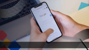 Seseorang sedang membuka halaman pencarian Google di smartphone, dengan laptop di latar belakang, menggambarkan aktivitas pencarian informasi yang berkaitan dengan optimasi website.