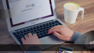 Seorang wanita menggunakan laptop MacBook Air di atas meja kayu, dengan layar menampilkan halaman pencarian Google. Di samping laptop terdapat secangkir kopi dan sebuah ponsel yang menampilkan aplikasi media sosial. Suasana kerja santai menggambarkan aktivitas digital marketing di Jakarta.