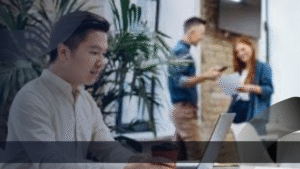 Seorang staf muda sedang bekerja dengan laptop di ruang kerja terbuka agensi periklanan, sementara dua rekan berdiskusi sambil memegang dokumen di latar belakang.