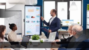 Seorang pria berpakaian formal sedang mempresentasikan data dan grafik di layar besar di ruang rapat modern, dikelilingi oleh tim kerja yang memperhatikan dengan serius. Presentasi ini menunjukkan pentingnya analisis produk dan strategi pemasaran—ilustrasi yang menggambarkan mengapa perusahaan butuh Product Specialist untuk mengelola pengembangan produk secara efektif.