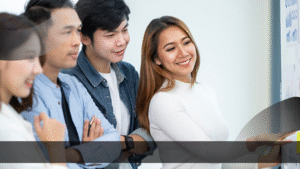 Sekelompok profesional muda tersenyum dan berdiskusi di depan papan berisi grafik dan data, dipandu oleh seorang wanita yang menunjuk pada presentasi visual. Suasana kerja tim yang kolaboratif ini menggambarkan pentingnya peran dalam analisis dan pengembangan produk—sebuah ilustrasi kuat bahwa perusahaan butuh Product Specialist untuk memimpin strategi produk yang sukses.