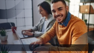 Marketing specialist muda sedang bekerja dengan laptop di kantor modern, tersenyum sambil berkolaborasi dengan rekan kerja.