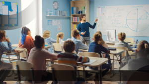 Seorang guru pria sedang menjelaskan konsep matematika dan geometri di papan tulis putih kepada sekelompok siswa sekolah dasar yang duduk rapi di dalam kelas yang terang dan modern.