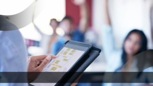 Tampilan close-up seseorang yang memegang tablet berisi bagan strategi dan rencana pembelajaran digital di tengah suasana ruang kelas atau diskusi yang interaktif