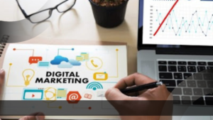 Tampilan close-up tangan seorang profesional sedang menulis rencana strategi di atas buku catatan bertuliskan 'DIGITAL MARKETING' yang dilengkapi berbagai ikon komunikasi. Di sampingnya, terdapat laptop yang menampilkan grafik performa data analitik.